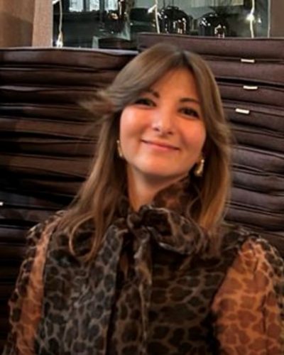 Smiling woman with shoulder-length brown hair wearing a leopard-print blouse with a bow, posing indoors.